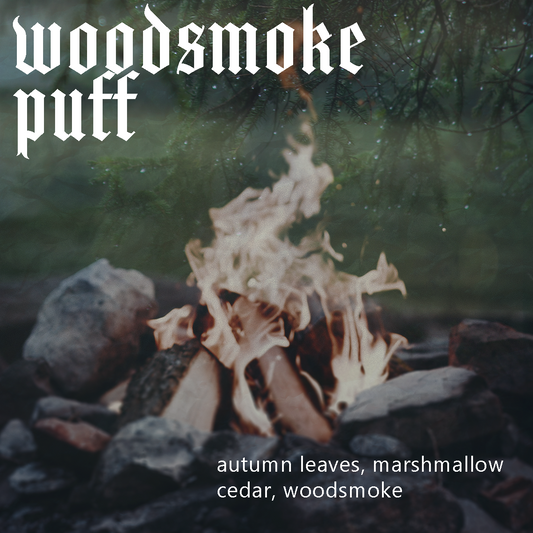 An image of a campfire burning outdoors with rocks and logs, overlaid with a transparent gradient image of raindrops on a conifer, the fragrance name text 'Woodsmoke Puff', and scent notes listed as 'autumn leaves, marshmallow cedar, woodsmoke' at the bottom right.